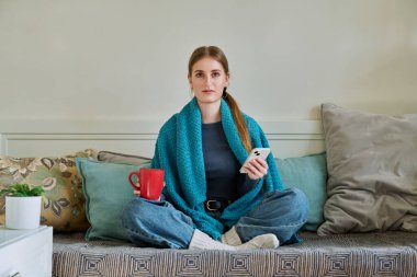 Sıcak kıyafetlerle battaniyenin altında akıllı telefonlu bir bardak sıcak içecekle evdeki kanepede oturan soğuk sezon, kışın tatilde hafta sonu. Yaşam tarzı rahatlık rahatlama gençlik konsepti