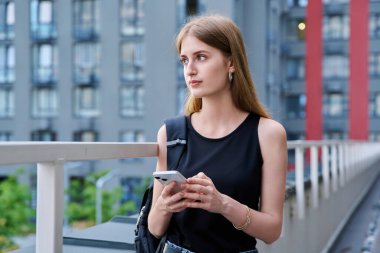 Akıllı telefon kullanan genç sarışın kadın, modern şehir geçmişi. Ergen öğrenci gezmek için mobil uygulama uygulamaları kullanarak mesajlaşıyor. Teknoloji İnternet Eğitim Gençlik Usulü