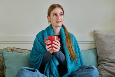 Soğuk mevsimde sıcak içecekle battaniyenin altında ısınan genç bir kadın, kışın tatilde evde koltukta oturuyor. Yaşam tarzı rahatlık rahatlama gençlik konsepti