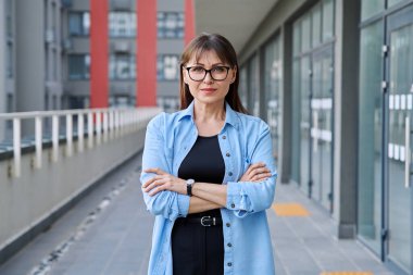 Açık havada gülümseyen, kendine güvenen olgun bir kadının şehir arka planında çekilmiş bir portresi. Yaşam tarzı, orta yaş, iş çalışma konsepti