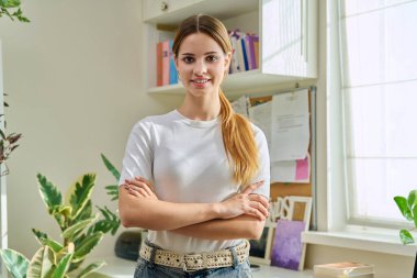 17-18 yaşlarındaki genç bir kızın portresi. Evde çapraz kollarla kameraya bakarak kendinden emin gülümsüyor. Uzun kızıl saçlı güzel bir kız. Gençlik yaşam tarzı güzellik modası
