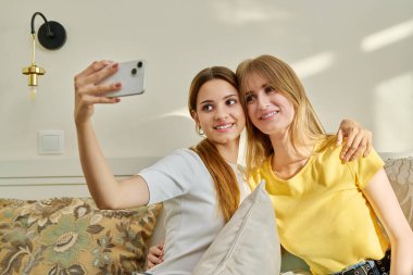 İki neşeli, pozitif, genç kız arkadaş evlerinde kanepede oturup akıllı telefondan selfie çekiyorlar. Gençler, gençler eğleniyor, arkadaşlık yaşam tarzı konsepti