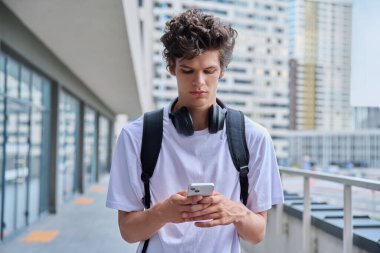 Genç adam akıllı telefon kullanıyor, modern şehir gökdelenleri caddesi. 20 yaşındaki yakışıklı erkek öğrenci telefon ekranına bakıyor. Gençlik, teknoloji mobil uygulamaları, internet iletişim hizmetleri