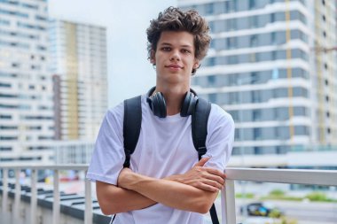 Dışarıdaki kameraya bakan yakışıklı genç adam, modern gökdelenler şehir tarzı arka plan. Kollarını kavuşturup sırt çantasına kıvırcık saçlar takmış 19,20 yaşında mutlu bir erkek öğrenci. Gençlik yaşam tarzı eğitimi