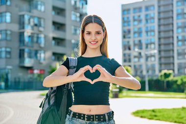 Young happy teenage girl student with backpack showing heart with hands, city background, urban style. Youth, happiness, love, joy, positive emotions, success concept clipart