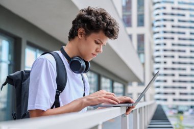 Genç erkek, dizüstü bilgisayar kullanıyor, modern şehir gökdelenleri geçmişi var. Yakışıklı bir öğrenci, 19-20 yaşlarında, boş zamanlarında bilgisayar kullanıyor, okuyor, çalışıyor. Teknoloji, gençlik, eğitim, kentsel tarz kavramı