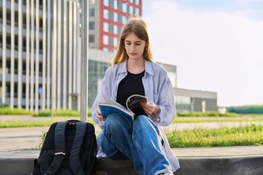 Genç bir üniversite öğrencisi, sırt çantalı güzel bir kız ders kitabı okuyor sınav sınavlarına hazırlanıyor, açık hava, modern şehir geçmişi. Gençlik, yaşam tarzı, eğitim kavramı
