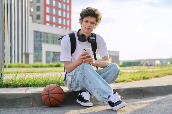 stock image Portrait of 19, 20 year old smiling student guy looking at camera, outdoors. Handsome curly young male with backpack, headphones, holding smartphone, sitting on floor with basketball ball. Youth