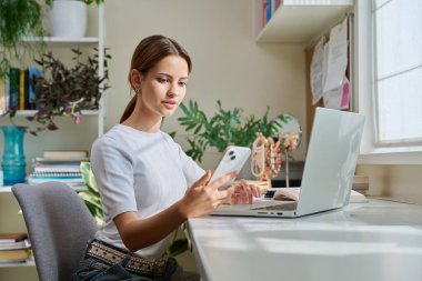 Ergen bir kız, 17 yaşında, 18 yaşında. Masasında dizüstü bilgisayarla oturuyor. Elinde akıllı telefon var. Eğitim, yaşam tarzı, gençlik konsepti