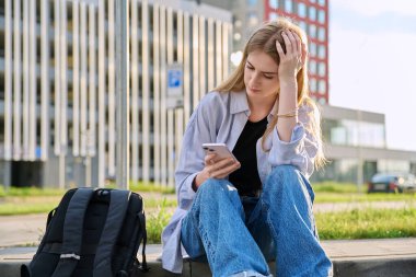 Üzgün, mutsuz, genç, şehir dışı tarzı bir bayan. Sırt çantalı kız üniversite öğrencisi akıllı telefon kullanıyor. Sorunlar, zorluklar, depresyon, gençlerin zihinsel sağlığı