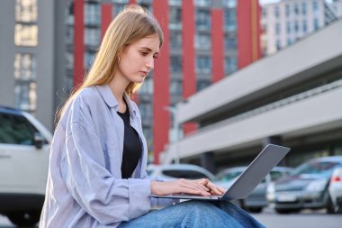 Genç, çekici, ciddi bir üniversite öğrencisi dışarıda dizüstü bilgisayar kullanıyor, modern şehir geçmişi var. Eğitim, teknoloji, eğitim, 17-18 yaş gençlik kavramı