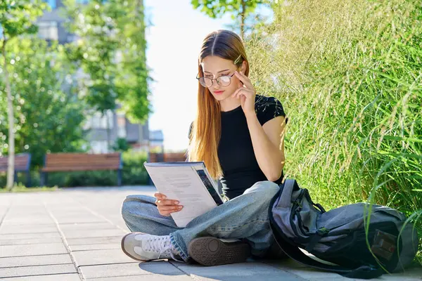 stock image Portrait of teenage student girl 17,18 years old sitting outdoors on floor with textbook backpack preparing for test exam. Education, youth, teenagers, college age, learning concept