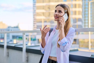17-19 yaşlarında güzel genç bir bayan cep telefonuyla konuşuyor. Gökdelenlerin arka planında modern bir şehir. Gençlik, yaşam tarzı, şehir tarzı konsepti