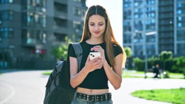 Sırt çantalı neşeli genç bayan dışarıda akıllı telefon kullanıyor. Gezici uygulamalar kullanarak ders çalışmak için mesajlaşan gülen genç kız öğrenci. Teknoloji İnternet Eğitim Gençliği