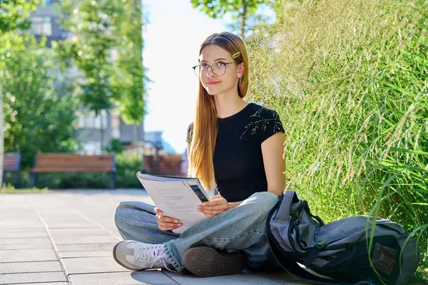 stock image Portrait of teenage student girl 17,18 years old sitting outdoors on floor with textbook backpack preparing for test exam. Education, youth, teenagers, college age, learning concept