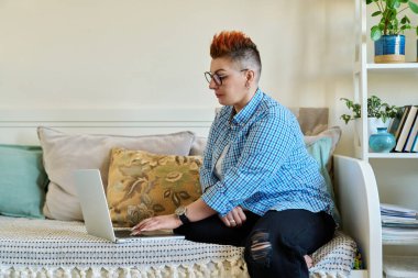 Evdeki koltukta dizüstü bilgisayar kullanan orta yaşlı bir kadın. Ciddi bir kadın, kırmızı saç kesimi, günlük giysiler içinde bilgisayarda çalışıyor, rahatlıyor, iletişim kuruyor.