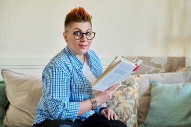 Evdeki kanepede oturan orta yaşlı bir kadın kitap okuyor. Boş zaman, hobi, okuma, kurgu, iş, eğitim, edebiyat.