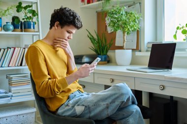 Evdeki akıllı telefondan haber okuyan mutsuz genç bir erkek. Üzgün adam üniversite öğrencisi haberleri okuyor. Sorunlar, depresyon, gençlerin akıl sağlığı