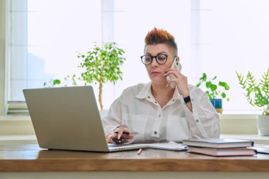 Orta yaşlı bir iş kadını cep telefonuyla konuşuyor. Masasında dizüstü bilgisayarla oturuyor. İş belgeleriyle çalışan kendine güvenen olgun bir kadın. İş, işyeri, 40 'lı yaşlarda insanlar.