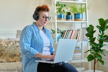 Kulaklıklı orta yaşlı bir kadın evdeki koltukta dizüstü bilgisayar kullanarak oturuyor. Ciddi bir kadın, kırmızı saçlı, günlük kıyafetleriyle bilgisayarda çalışıyor, rahatlıyor, iletişim kuruyor.