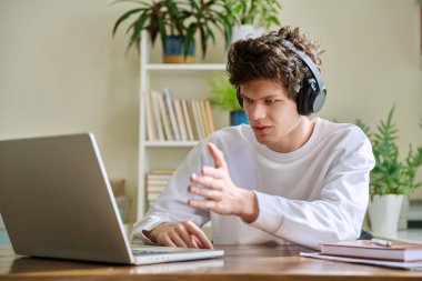 Kulaklıklı genç bir üniversite öğrencisi web kamerası bilgisayarına bakıyor, konuşuyor, internetten ders çalışıyor, evinde oturuyor. E-öğrenme, uzaktan eğitim, eğitim, gençlik, teknoloji kavramı