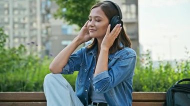 Kulaklıklı, müzik dinleyen, dışarıda bankta rahatça oturan mutlu genç bir kadın. Olumlu, neşeli, gözleri kapalı bir kadının portresi. Gençlik, teknoloji, yaşam tarzı, duygular