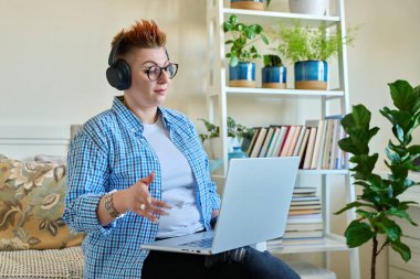 Kulaklıklı orta yaşlı kadın evdeki kanepede oturan dizüstü bilgisayarını kullanıyor. 40 'lı yaşlarda kadın ekrandaki görüntülü sohbet konferansına bakıyor online toplantı eğitim danışmanlığı teknolojisi