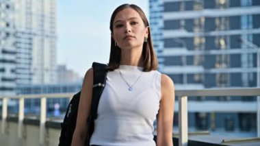 Dışarıdaki kameraya bakan kendine güvenen genç bir kız öğrencinin portresi. Çapraz kollu ciddi bir kadın, şehir çağdaş gökdelenleri arka planı, şehir tarzı. Gençlik, yaşam tarzı konsepti