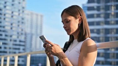 Sırt çantalı güzel bir kadın dışarıda akıllı telefon kullanıyor, profil manzaralı. Şehir tarzı mesaj yazan neşeli kadın mesajlaşması. Teknoloji mobil uygulamaları İnternet çevrimiçi yaşam tarzı