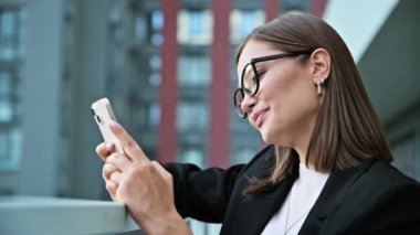 Akıllı telefon kullanan başarılı genç bir iş kadını, açık ofis binası geçmişi. Gülümseyen olumlu kadın mesajları okuyor. Teknoloji sektörü girişimciliği gençlik kavramı