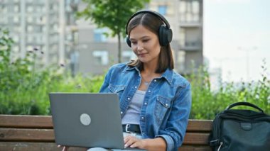 Kulaklıklı genç ve ciddi bir bayan öğrenci dışarıda bankta dizüstü bilgisayara bakıyor. Eğitim teknolojileri boş vakitlerde iletişim sağlar. Uzak ders, webinar, online toplantı, gençlik