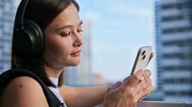 Sırt çantalı, kulaklıklı, açık havada akıllı telefon kullanan, profil manzaralı güzel bir kadın. Şehir tarzı mesaj yazan neşeli kadın mesajlaşması. Teknoloji mobil uygulamaları yaşam tarzı