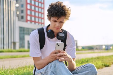 Modern şehir sokaklarında akıllı telefon kullanan genç bir adam. 20 yaşındaki yakışıklı erkek öğrenci telefon ekranına bakıyor. Gençlik, teknoloji mobil uygulamaları, internet iletişim hizmetleri, yaşam tarzı