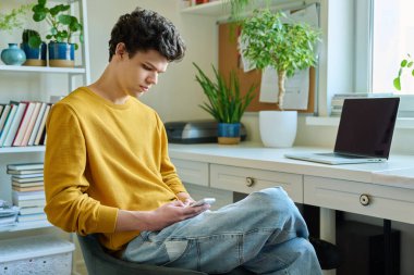 Genç adam akıllı telefon kullanıyor, evde oturuyor. Yakışıklı üniversite öğrencisi 19-20 yaşlarında, sarı saçlı, kıvırcık saçlı. Teknolojiler, mobil uygulamalar, iletişim eğlence kavramı