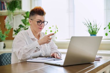 Orta yaşlı iş kadını ev ofisteki masada oturan dizüstü bilgisayarında görüntülü sohbet görüşmesi yapıyor. Çevrimiçi iletişim, iş teknolojisi, girişimcilik işi, danışmanlık eğitimi