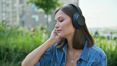 Kulaklıklı, müzik dinleyen, dışarıda bankta rahatça oturan mutlu genç bir kadın. Olumlu, neşeli, gözleri kapalı bir kadının portresi. Gençlik, teknoloji, yaşam tarzı, duygular