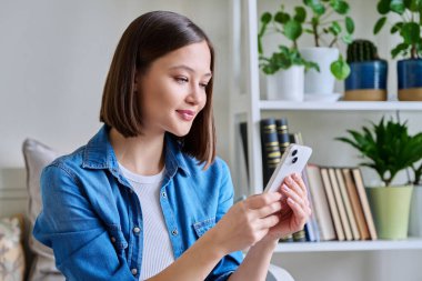Evdeki koltukta oturan akıllı telefonu kullanan genç bir kadın. Kadın mesajlaşma görüntüsü, mobil uygulamalar teknoloji iş eğitimi için internet hizmetleri eğlence iletişim alışveriş yaşam tarzı