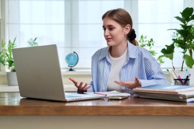 Genç bir kız öğrenci masasında oturmuş bilgisayar kamerasıyla konuşuyor. 17 yaşında bir kız uzaktan eğitim görüyor, görüntülü konferans görüşmesi yapıyor, e-öğrenim teknolojisi eğitimi alıyor.