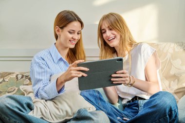 İki genç kız arkadaş evde kolej öğrencileri dijital tablete bakıp kanepede oturuyorlar. Eğitim teknolojisi, gençlik arkadaşlık kavramı.