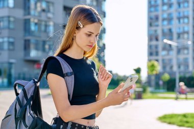 Sırt çantalı, akıllı telefon kullanan, modern şehir geçmişi olan genç ve ciddi bir bayan. Öğrenci gezisi için mobil uygulama uygulamaları kullanarak mesajlaşıyor. Teknoloji İnternet Eğitimi