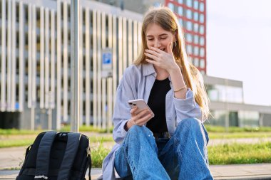 Genç gülen neşeli sarışın kadın akıllı telefon kullanıyor, modern şehir geçmişi. Öğrenci boş zamanlarında cep telefonu uygulamaları kullanarak mesajlaşıyor. Teknoloji eğitimi gençlik tarzı