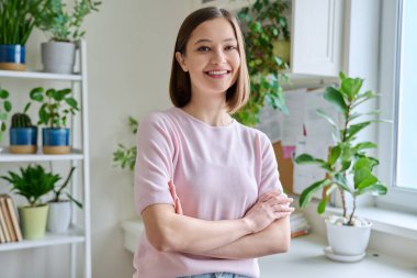 Evde kollarını kavuşturarak gülümseyen genç bir kadının portresi. 20 'li yaşlarda güzel bir kadın kameraya bakıyor. Güzellik, gençlik, mutluluk, sağlık, yaşam tarzı konsepti