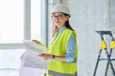 İnşaattaki kadın inşaatçı mimar müfettişin portresi. Yelek kasklı, kendinden emin orta yaşlı bir kadın kameraya bakıyor. İnşaat endüstrisi teknik meslek mühendisliği