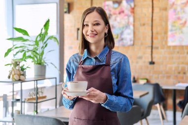 Başarılı genç bayan servis elemanı apronlu garson bir fincan kahveyle kafeteryanın içindeki kahvecinin kamerasına bakıyor. Küçük işletme işgali girişimciliği