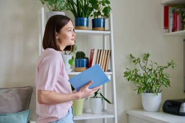 Mutlu gülümseyen, odaklanmış, pencerenin yanında kitap okuyan genç bir kadın. Önemli eğitim edebiyatı, ilginç kurgu, hobi kitapları.