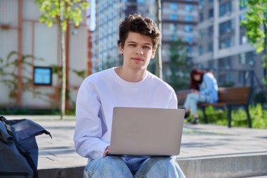 Genç yakışıklı üniversite öğrencisi dışarıda dizüstü bilgisayar kullanıyor, eğitim binası geçmişi var, kameraya bakan gülümseyen bir erkek. Eğitim, teknoloji, eğitim, 19-20 yaş gençlik kavramı
