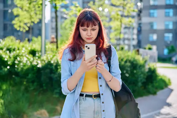 Sırt çantası açık havada akıllı telefon kullanan genç bir üniversite öğrencisi. Yüz piercingli kızıl saçlı, modern hippi kız. Moda, güzellik, eğitim, üniversite eğitimi, gençlik kavramı