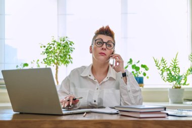 Orta yaşlı bir iş kadını cep telefonuyla konuşuyor. Masasında dizüstü bilgisayarla oturuyor. İş belgeleriyle çalışan kendine güvenen olgun bir kadın. İş, işyeri, 40 'lı yaşlarda insanlar.
