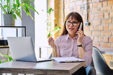 Orta yaşlı gülümseyen bir iş kadınının portresi dizüstü bilgisayarla çalışıyor, telefonla konuşuyor, ofis içinde masa başında oturuyor. İş, uzaktan kumanda, serbest çalışma, teknoloji, 40 'lı yaşlarda insanlar