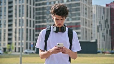 Açık havada akıllı telefon kullanan yakışıklı genç bir erkek. 19-20 yaşlarında, sırt çantalı kulaklıklı, yaşam tarzı, gençlik kavramı olan bir öğrenci. Eğitim, üniversite eğitimi, gençlik yaşam tarzı konsepti